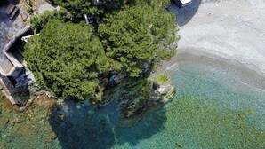 Vista aérea de una zona costera con playa despejada, un promontorio rocoso cubierto de vegetación densa y una pequeña estructura en la orilla.