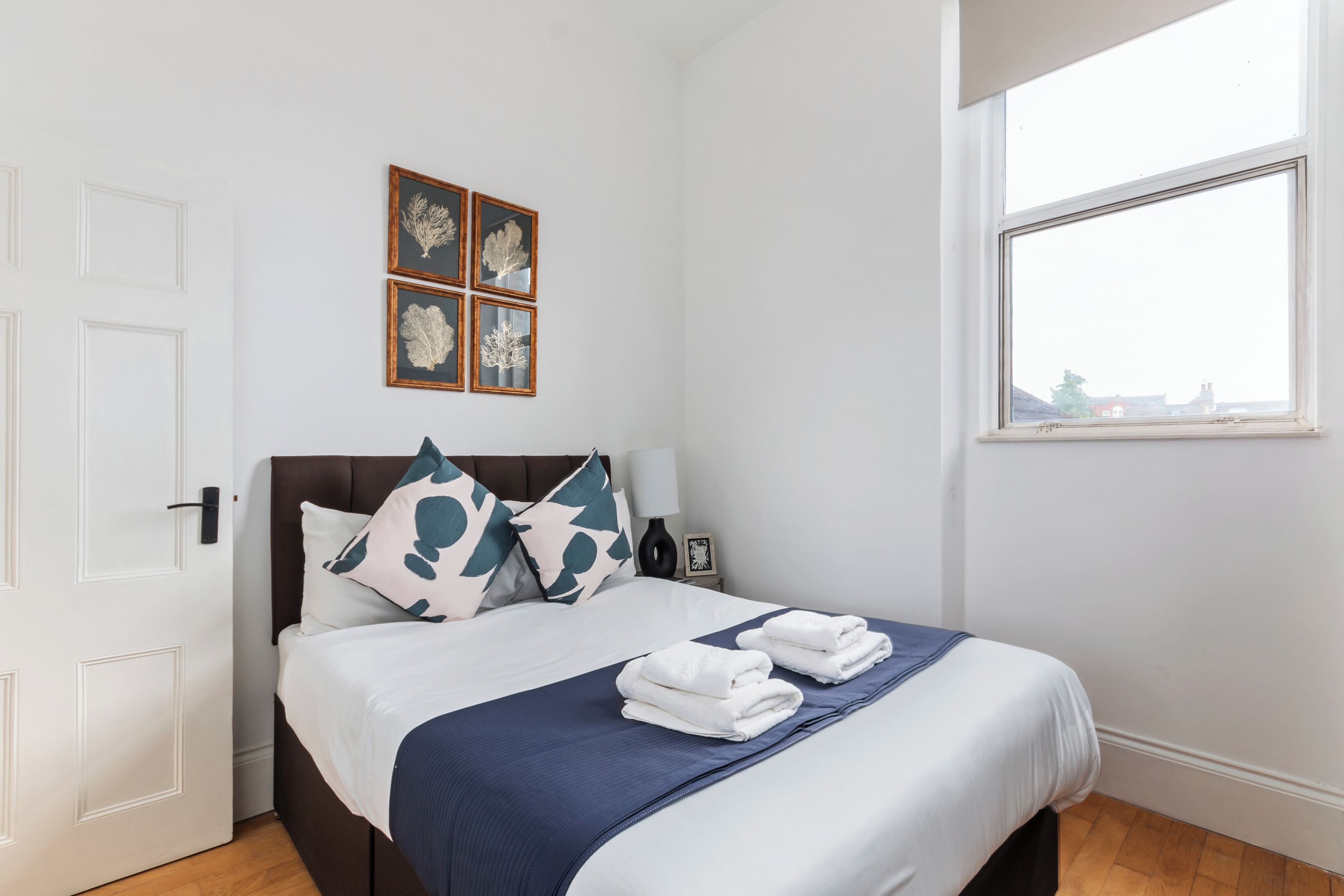 Een net opgemaakt bed met een blauw-witte spreidek, twee kussens en opgevouwen handdoeken op een houten vloer.