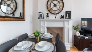 Un coin repas avec une cheminée blanche, deux grandes horloges murales et un téléviseur posé sur un meuble en bois.