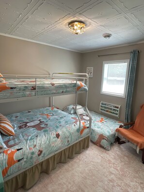 Une chambre avec un lit superposé, une couvre-lit à motifs floraux, un luminaire au plafond, une fenêtre avec des stores et une petite étagère avec des livres.