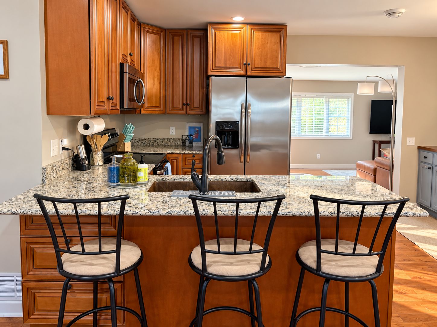 A kitchen with wooden cabinets, a stainless steel refrigerator, granite countertops, and three bar stools.