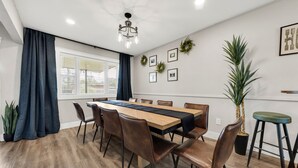 A modern dining room with a long wooden table, brown leather chairs, a chandelier, and a large potted plant. - A&M Getaway • Game Room • Mini Golf • Sleeps 12 (Bryan)