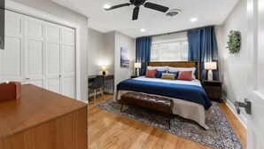 A bedroom with a bed, bedside tables, a desk, a chair, a ceiling fan, and a window with curtains. - A&M Getaway • Game Room • Mini Golf • Sleeps 12 (Bryan)