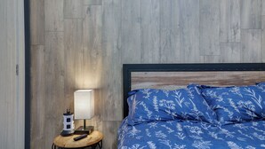Une chambre avec un mur en bois, un lit recouvert d’une couette à motifs floraux bleus, une table de chevet avec une lampe et une multiprise.