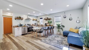 Une cuisine moderne avec un îlot central, des tabourets de bar et une table à manger. Le salon comprend un canapé et une grande fenêtre.