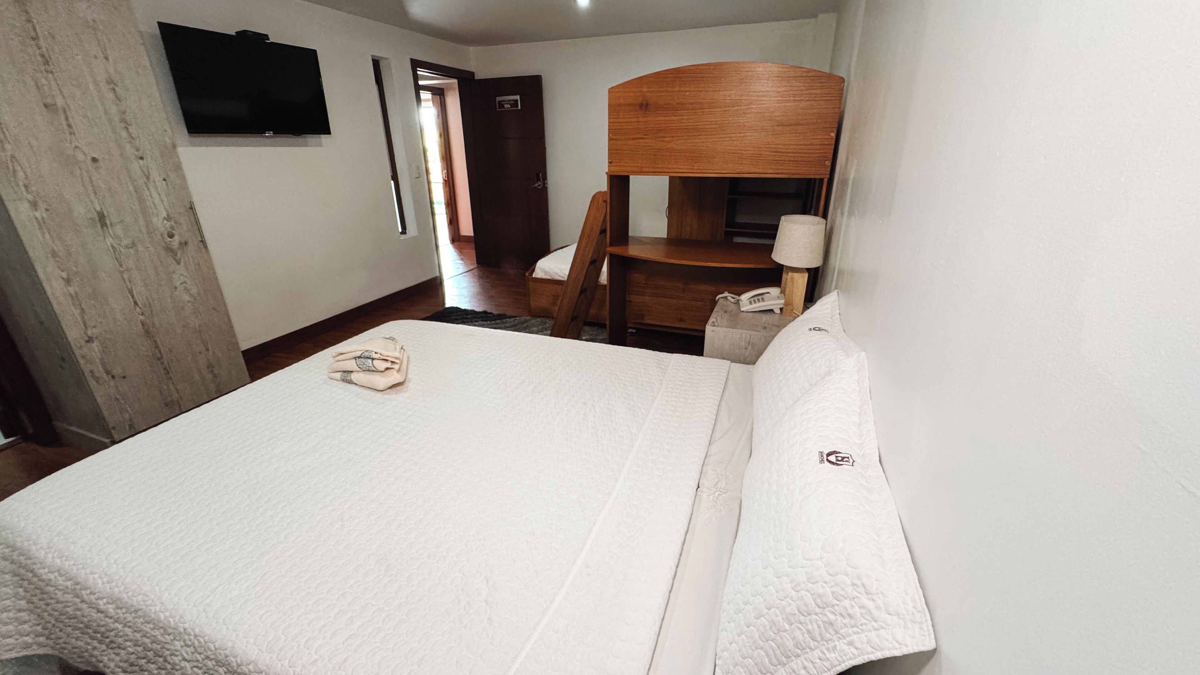 A hotel room with a bed, a wooden chair, a small table, and a television mounted on the wall.