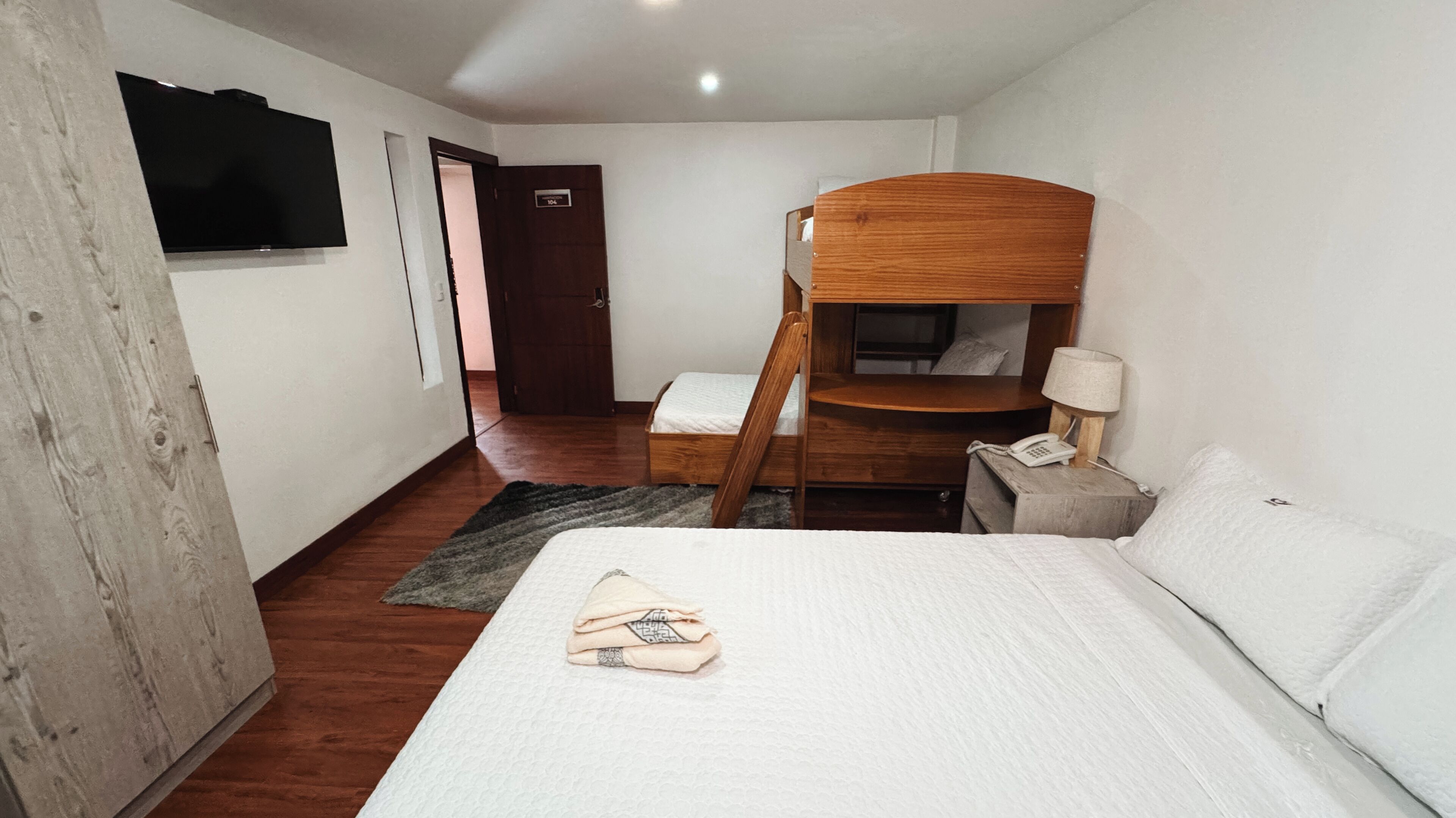 Ein Hotelzimmer mit einem Etagenbett, einem Schreibtisch mit Lampe, einem Telefon und einem an der Wand befestigten Fernseher.