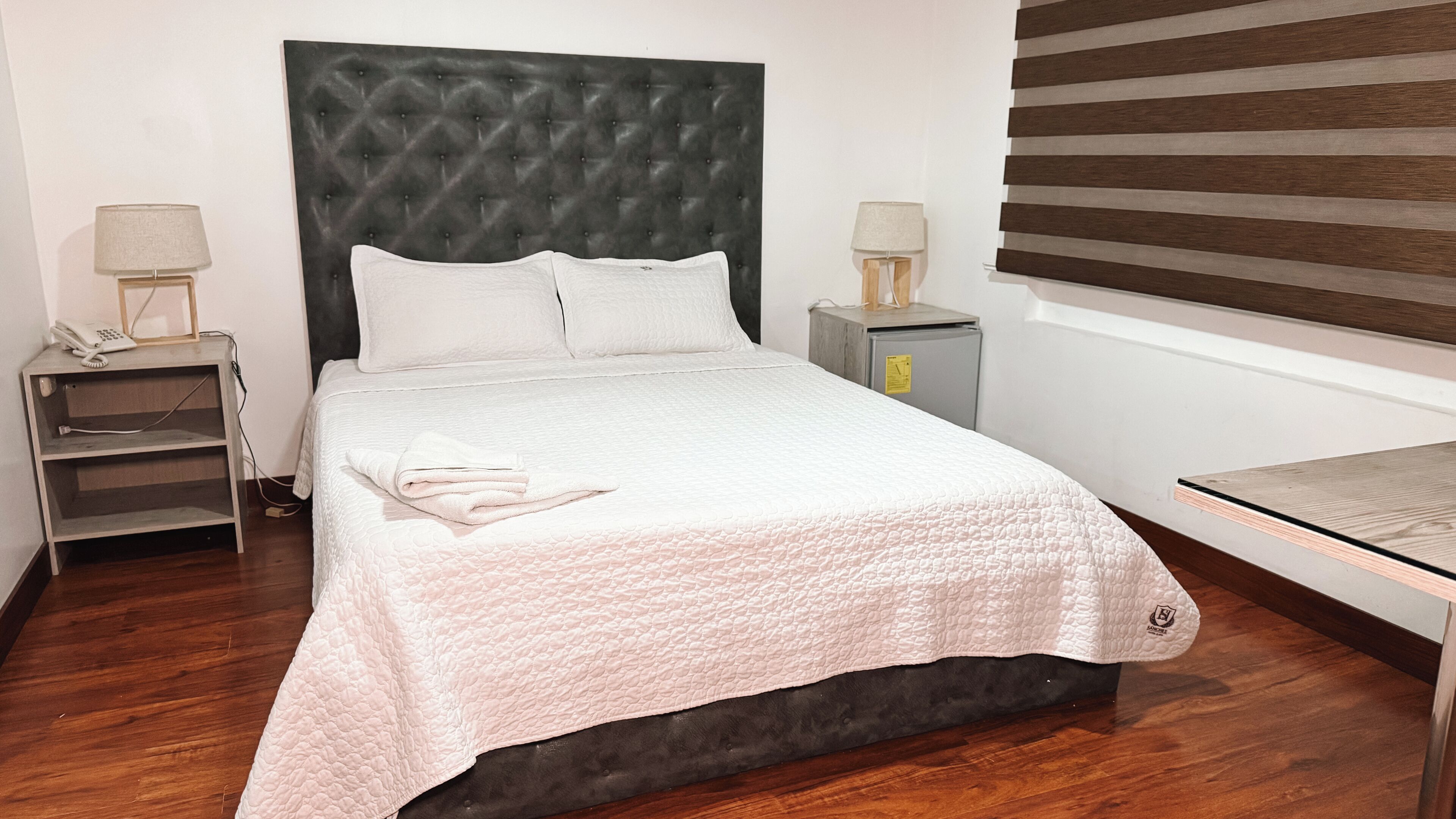 Una cama bien hecha con una colcha blanca acolchada, dos mesitas de noche con lámparas, un pequeño tocador y suelo de madera.