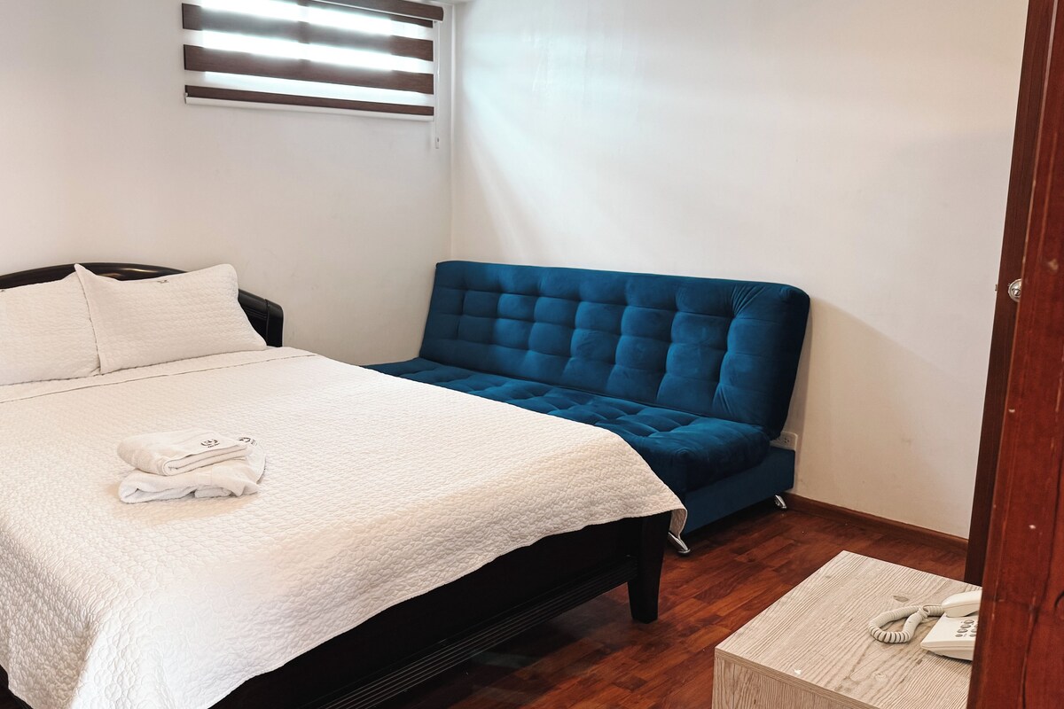 A neatly made bed with a white quilt and pillows, a blue upholstered headboard, and a wooden bedside table with a phone.