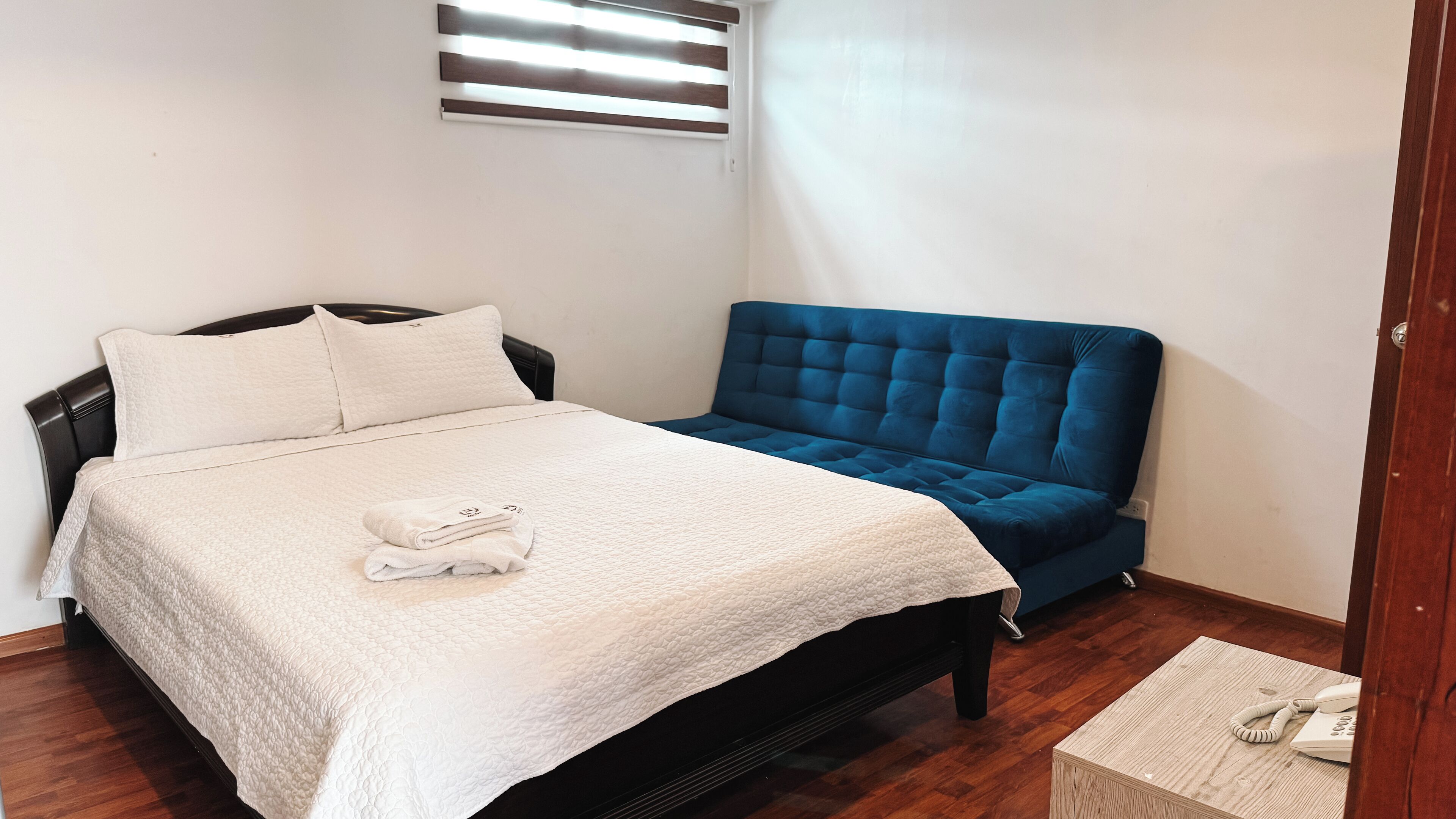 Un letto rifatto con coperta bianca e cuscini, una testiera imbottita blu e un comodino in legno con un telefono.