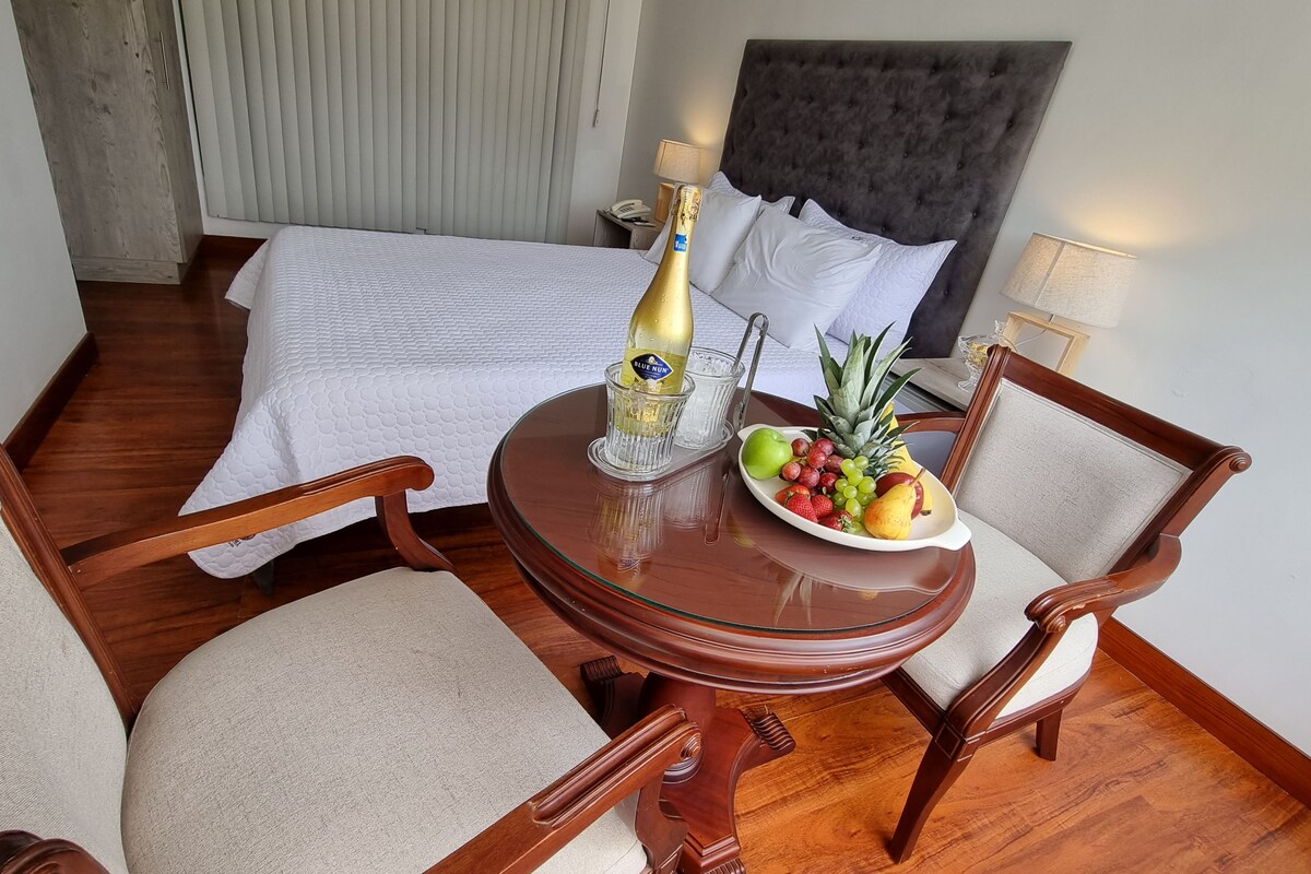 A hotel room with a bed, a small round table with a bottle of champagne, a fruit bowl, and two chairs.