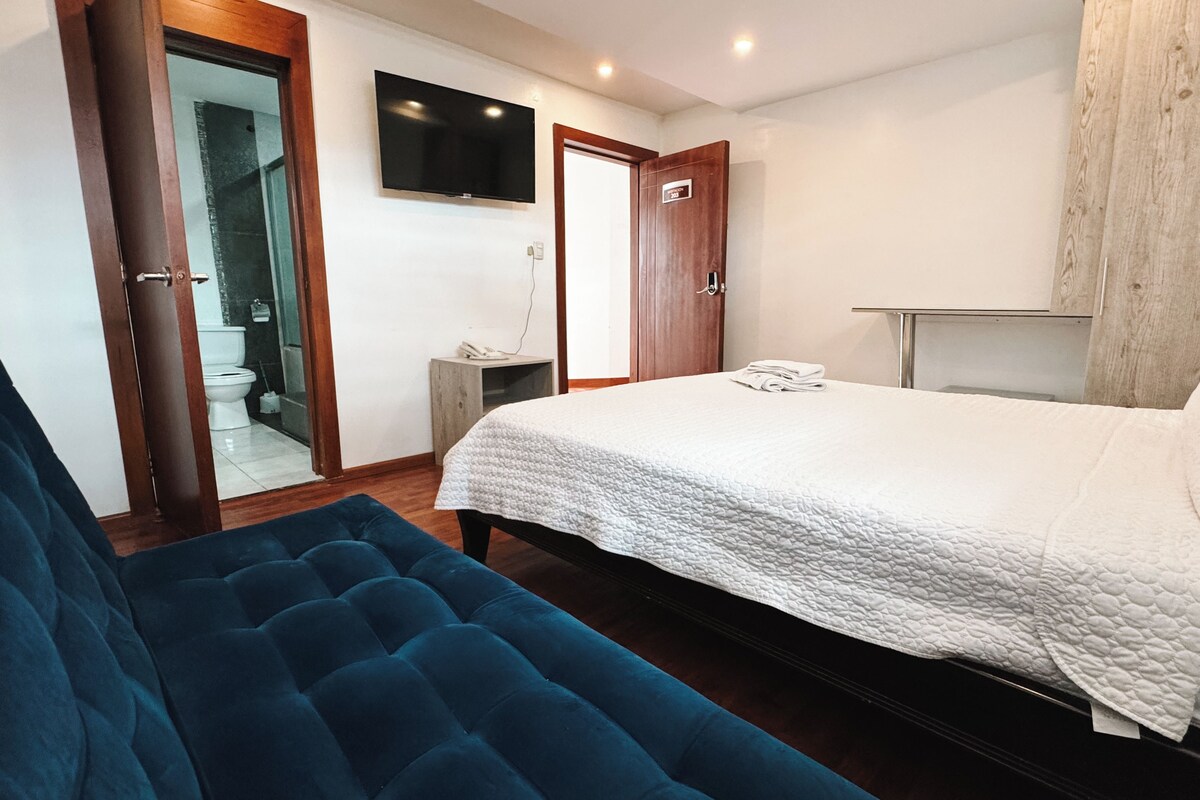 A bedroom with a bed, a blue sofa, a TV, a wooden door, and a small bedside table.