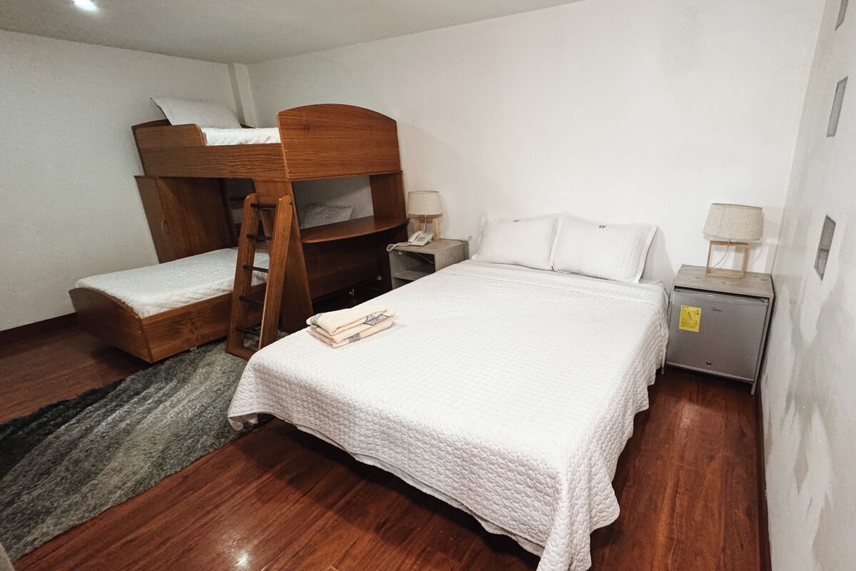 A bedroom with a bunk bed, a single bed, a bedside table with a lamp, and a small cabinet.