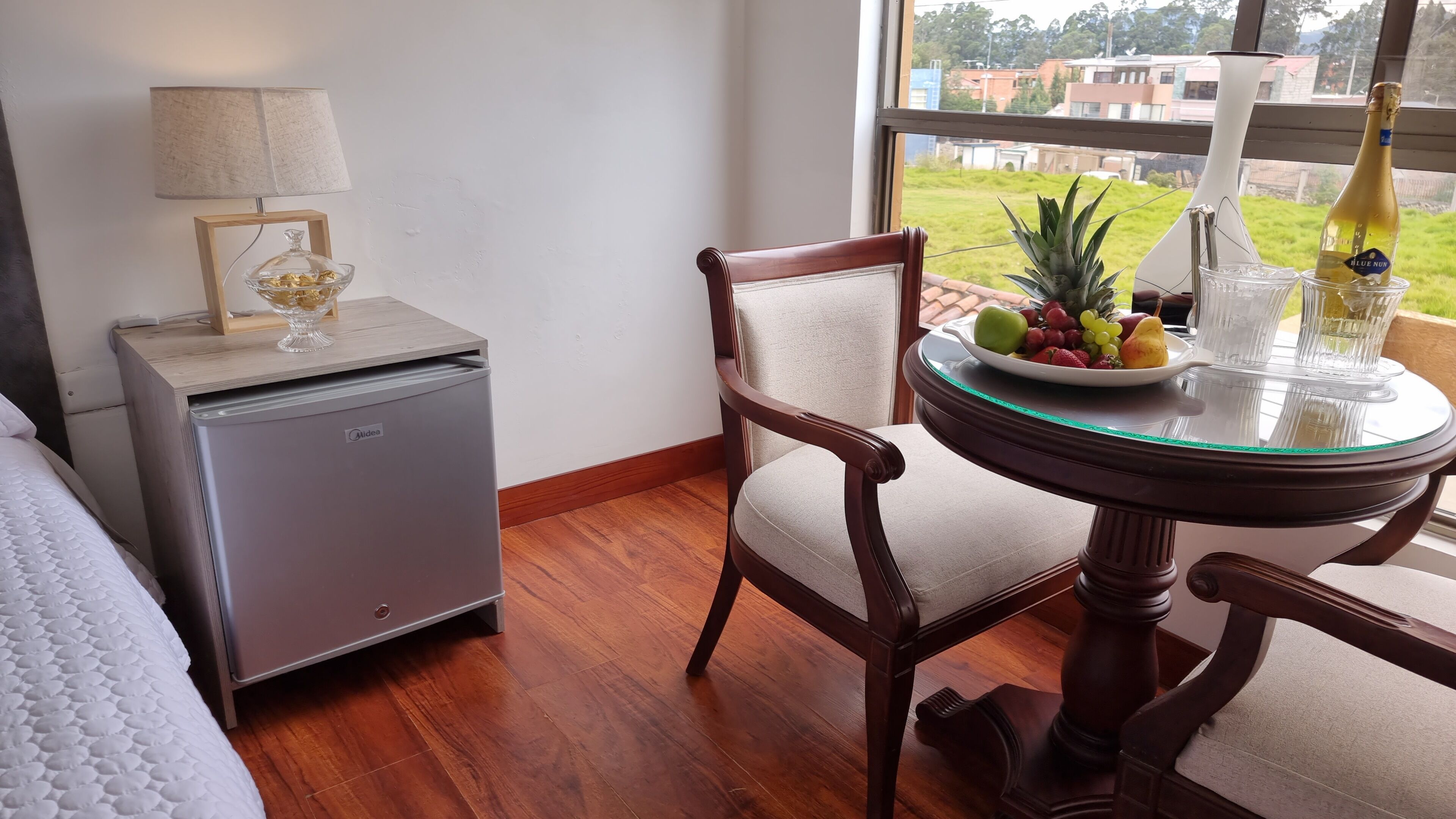 Ein Hotelzimmer mit Bett, Minibar, Esstisch mit Früchten und einer Flasche sowie Blick auf Gebäude durch ein Fenster.