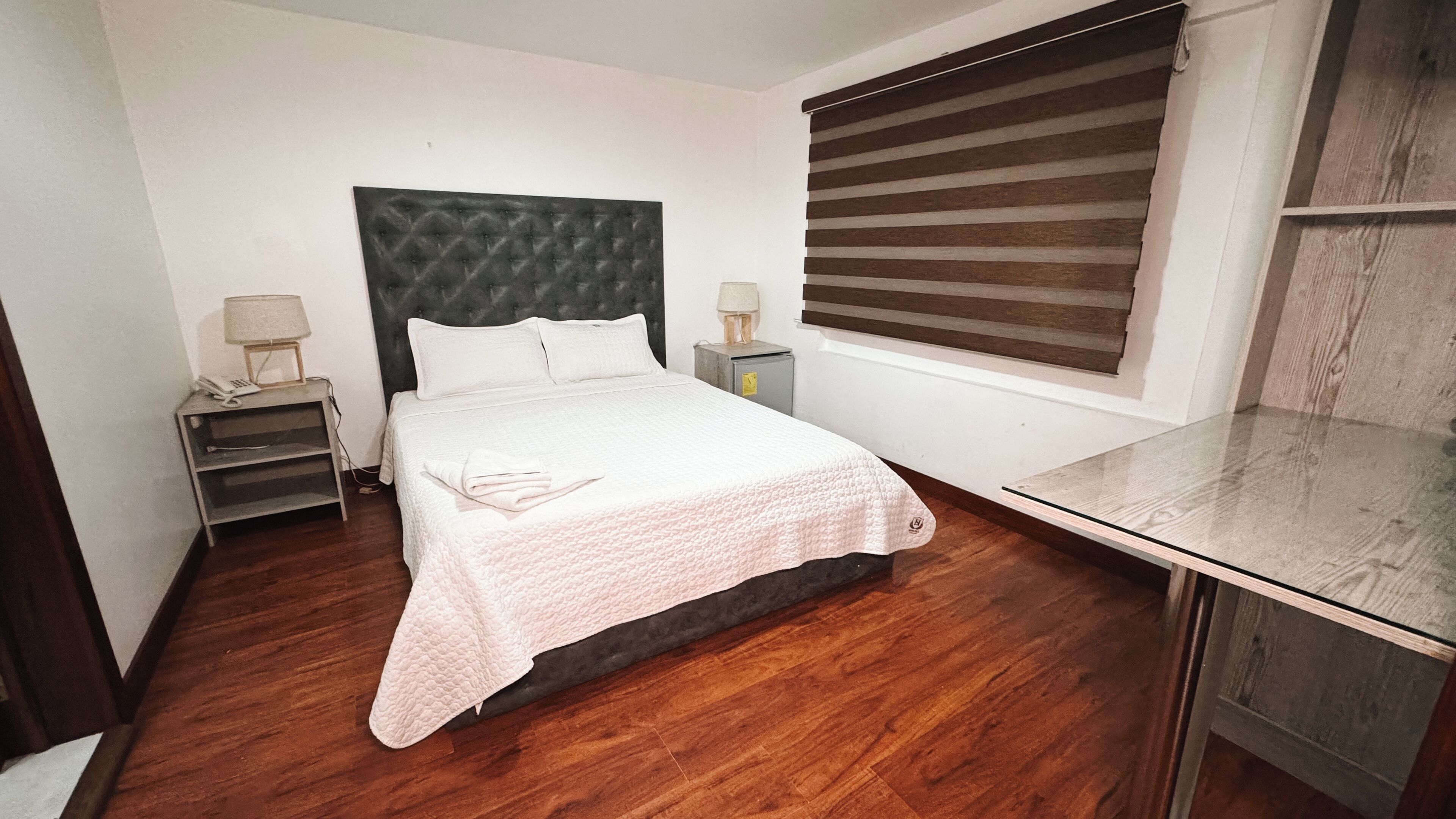 Un dormitorio con cama, lámparas de noche, mesita de noche, escritorio de madera y ventana con persianas.