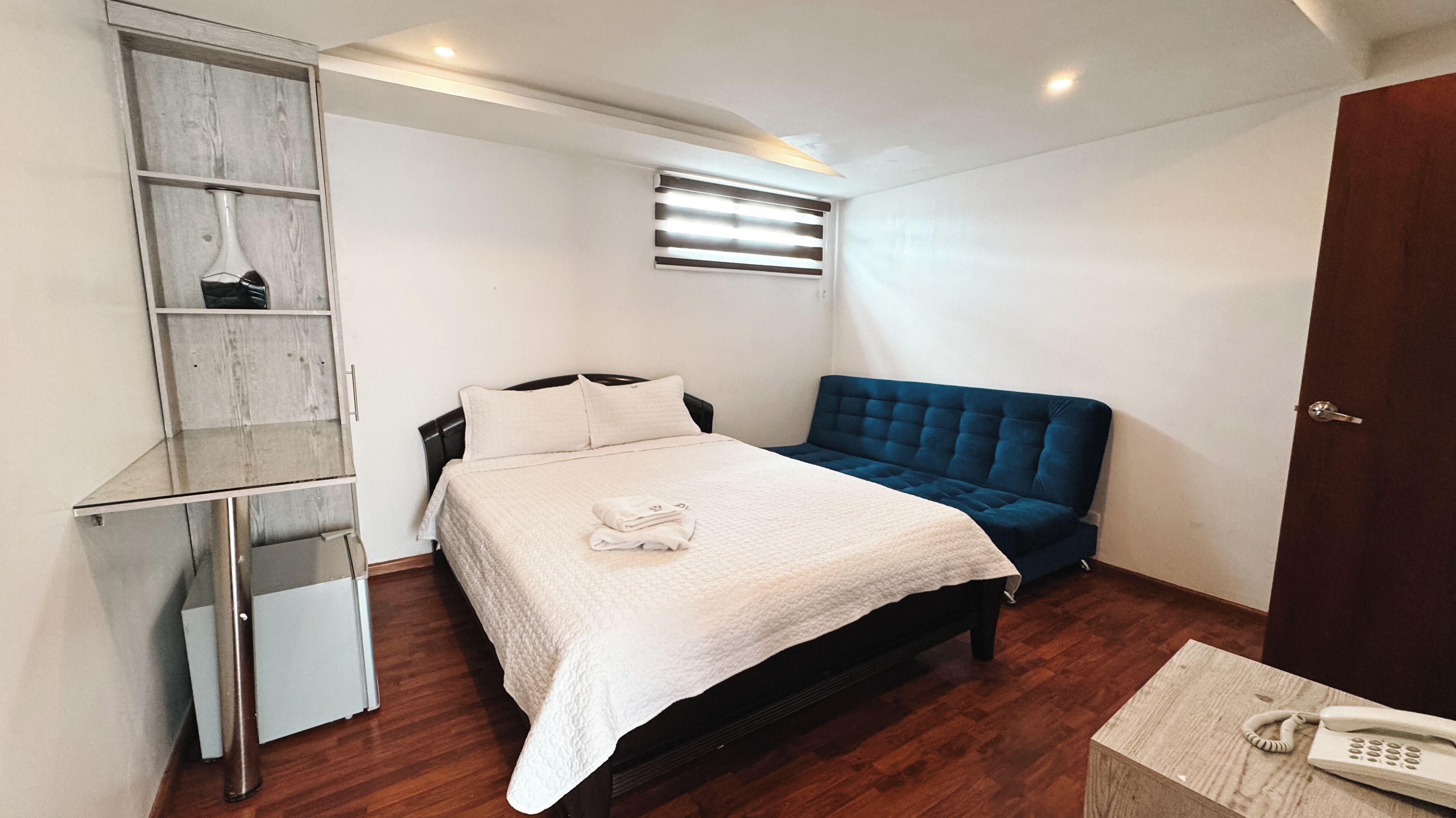 A bedroom with a bed, a blue upholstered chair, a bedside table with a telephone, and a wooden shelf unit.