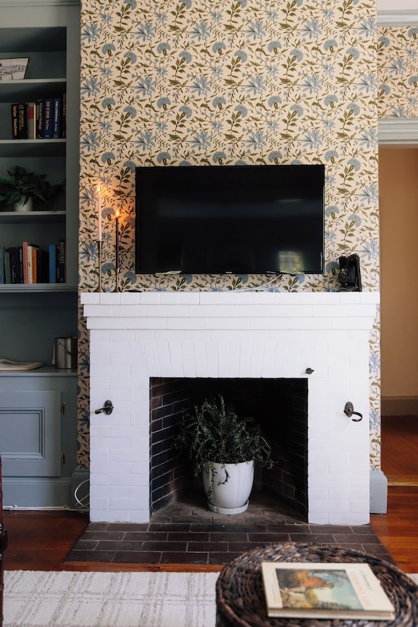 A fireplace with a TV above it, surrounded by patterned wallpaper, a bookshelf, and a potted plant.