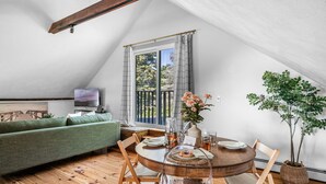 A cozy loft bedroom with a large window, wooden flooring, a green sofa, a dining table with chairs, and a TV on a stand. - The Ipswich Inn (Ipswich)