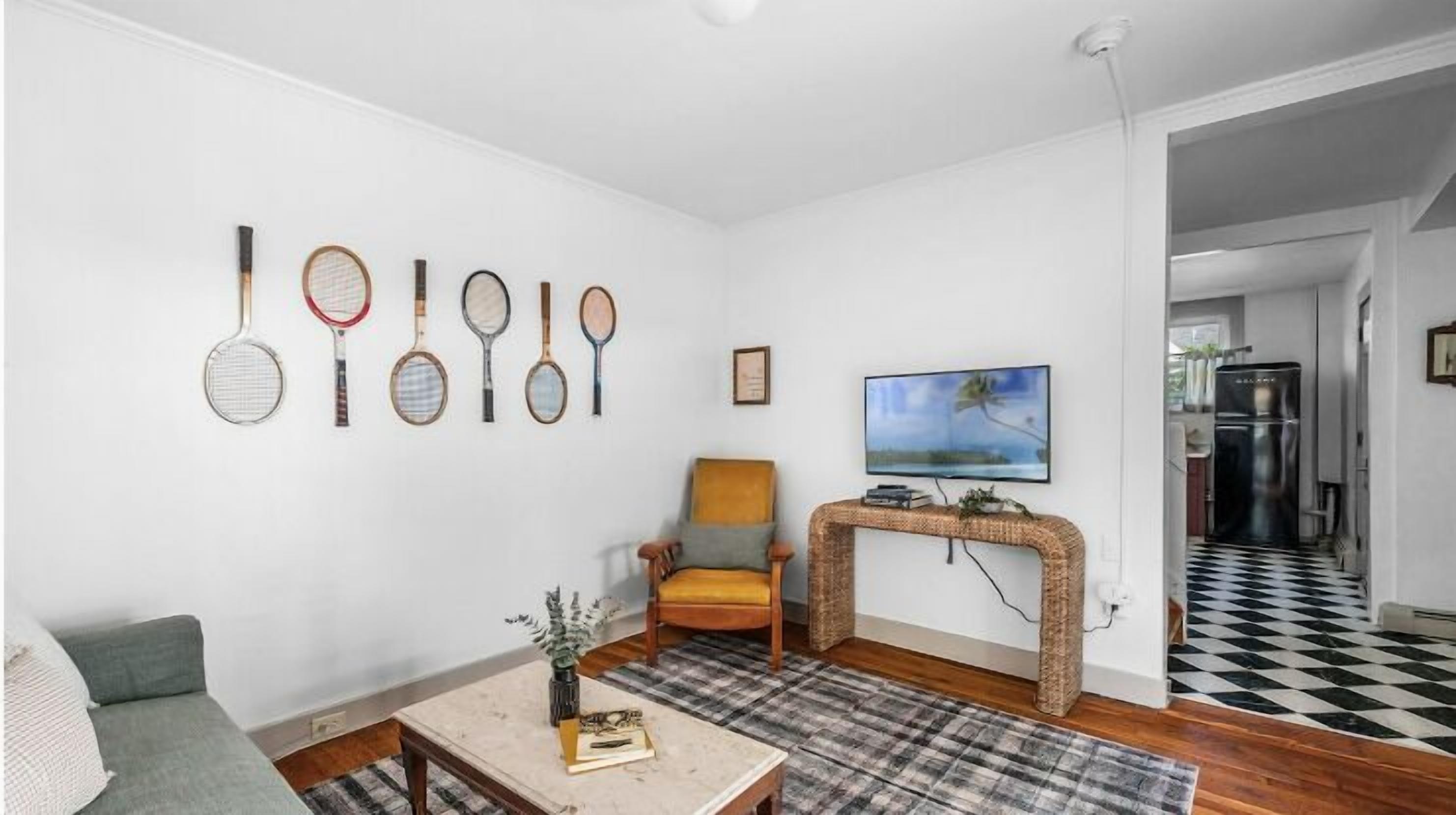 Un salon comprenant un canapé, un fauteuil, une table basse et un téléviseur fixé au mur. Plusieurs raquettes de tennis sont exposées sur le mur.
