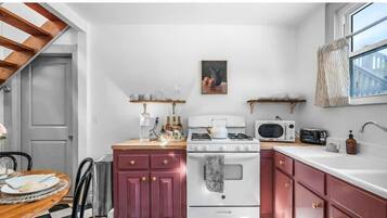 Una cocina compacta con armarios blancos y rosa, una estufa, microondas y un pequeño comedor con mesa y sillas.