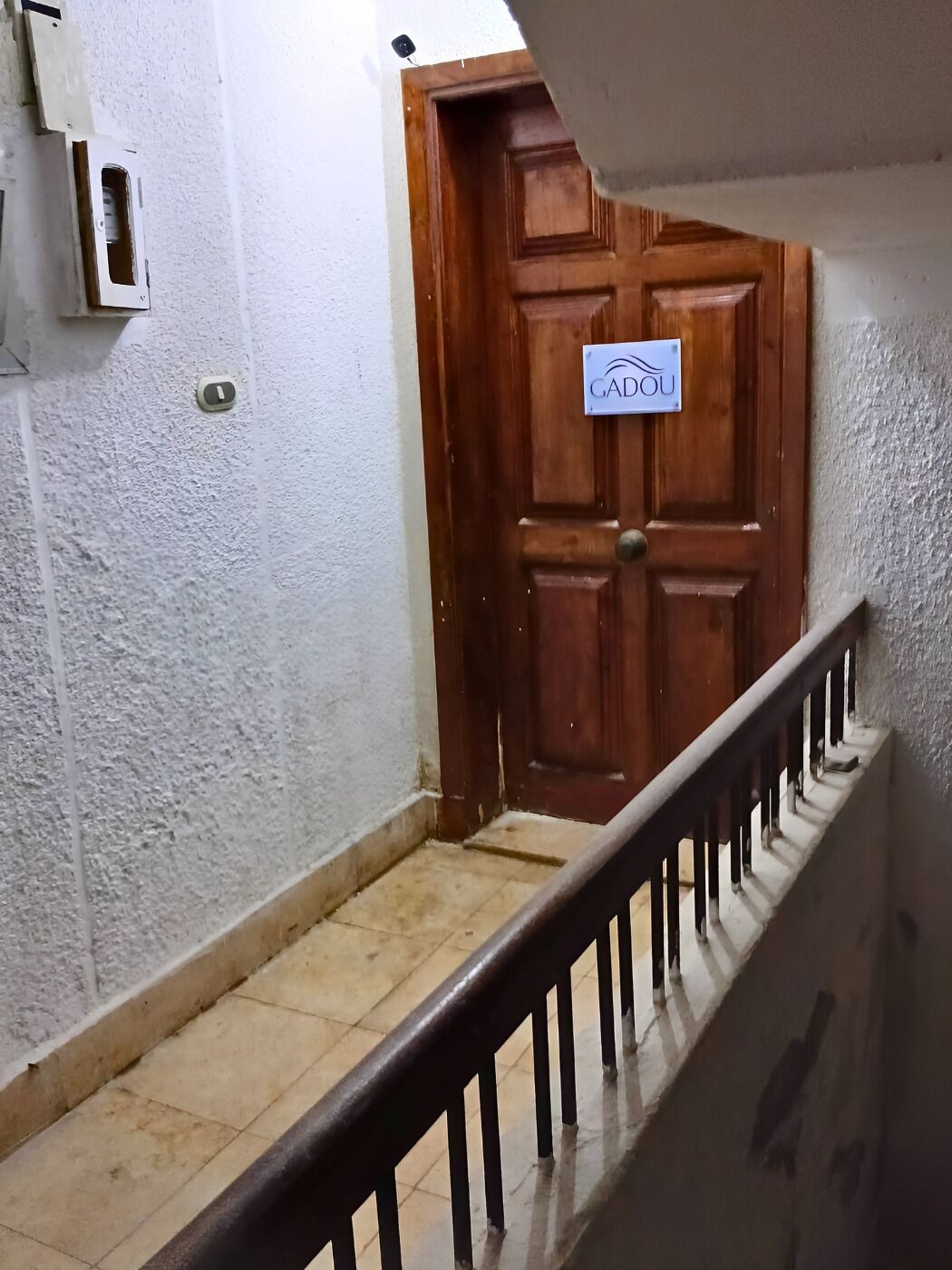 A wooden door with a sign reading "GADOU" and a metal railing leading to a tiled floor.