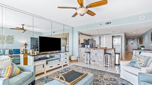 A modern living room with a large flat-screen TV, a ceiling fan, and a kitchen area visible through glass walls. - Destin Dreams 🌊 Gulf-View Condo at Sterling Shores Resort in Destin, Florida (Destin)