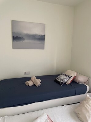 Een enkel bed met een donkerblauwe matras, twee opgerolde handdoeken, een kussen met een auto-opdruk en een kussen met een bloemenmotief.