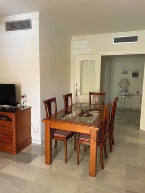 A dining area with a glass-top table surrounded by wooden chairs, a wooden cabinet with a television, and a door leading to another room. - Los Nidos Apartment, Carihuela (Torremolinos)