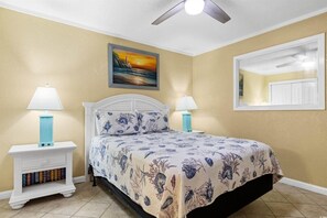 A bedroom with a bed, two bedside tables with lamps, a bookshelf, a ceiling fan, and a painting on the wall.