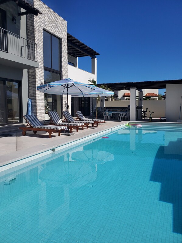 A swimming pool with lounge chairs and umbrellas, a modern building with a brick facade, and a clear blue sky.