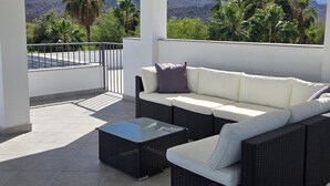 Een overdekt terras met een bank en salontafel, een plafondventilator en uitzicht op bergen en palmbomen.