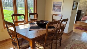 A dining area with a wooden table and chairs, a bowl of fruit, and a view of a garden through large windows.