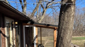 A wooden deck with a glass door leading to a covered area, a metal chair, and a tree.