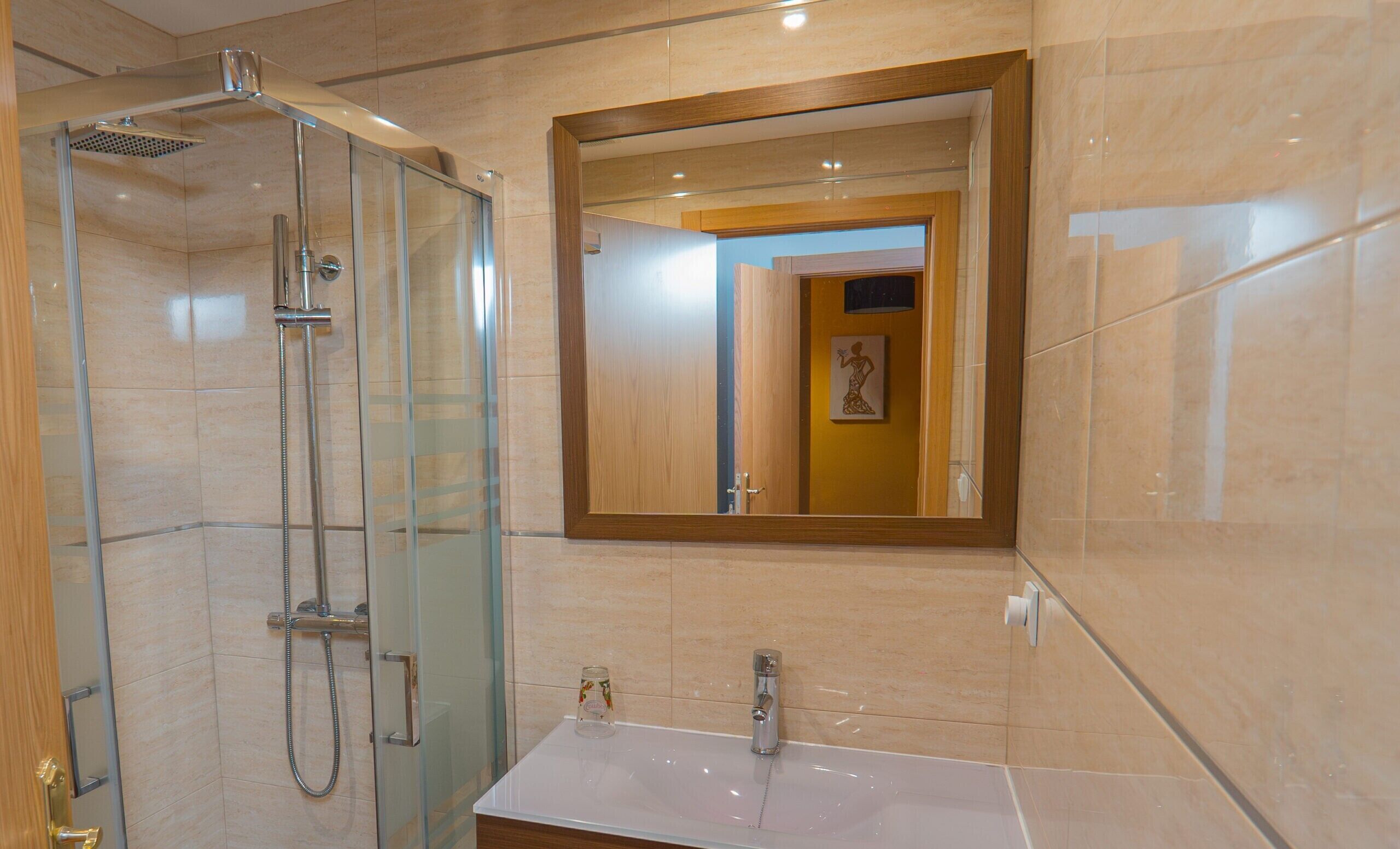 Un baño con ducha de vidrio, lavamanos, espejo y una puerta que da a otra habitación.