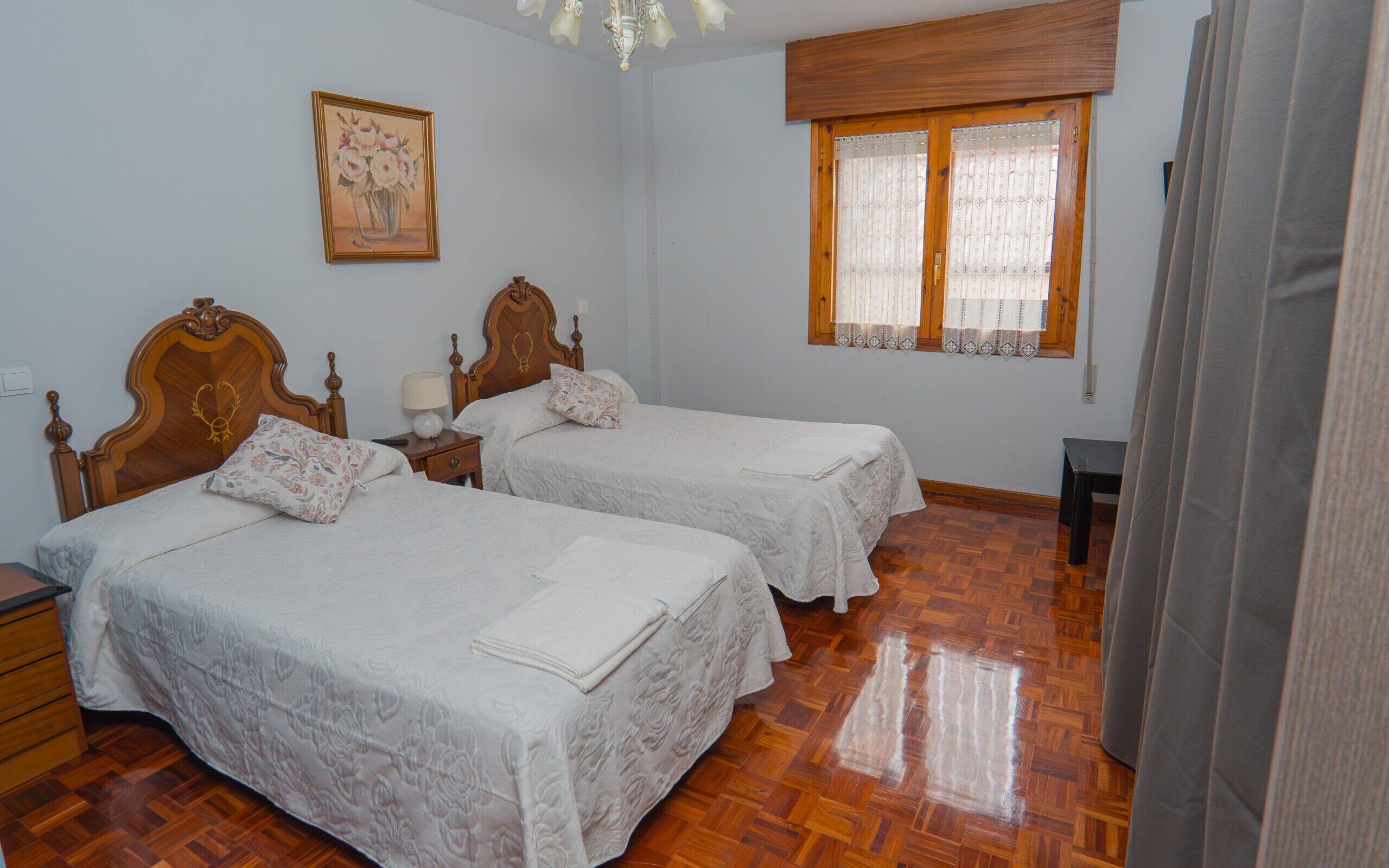 Una habitación con dos camas de madera, una mesita de noche con lámpara, cabecero de madera, una ventana con cortinas estampadas y suelo de madera.