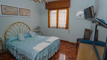 A bedroom with a bed, a desk, a chair, a television, and artwork on the wall.