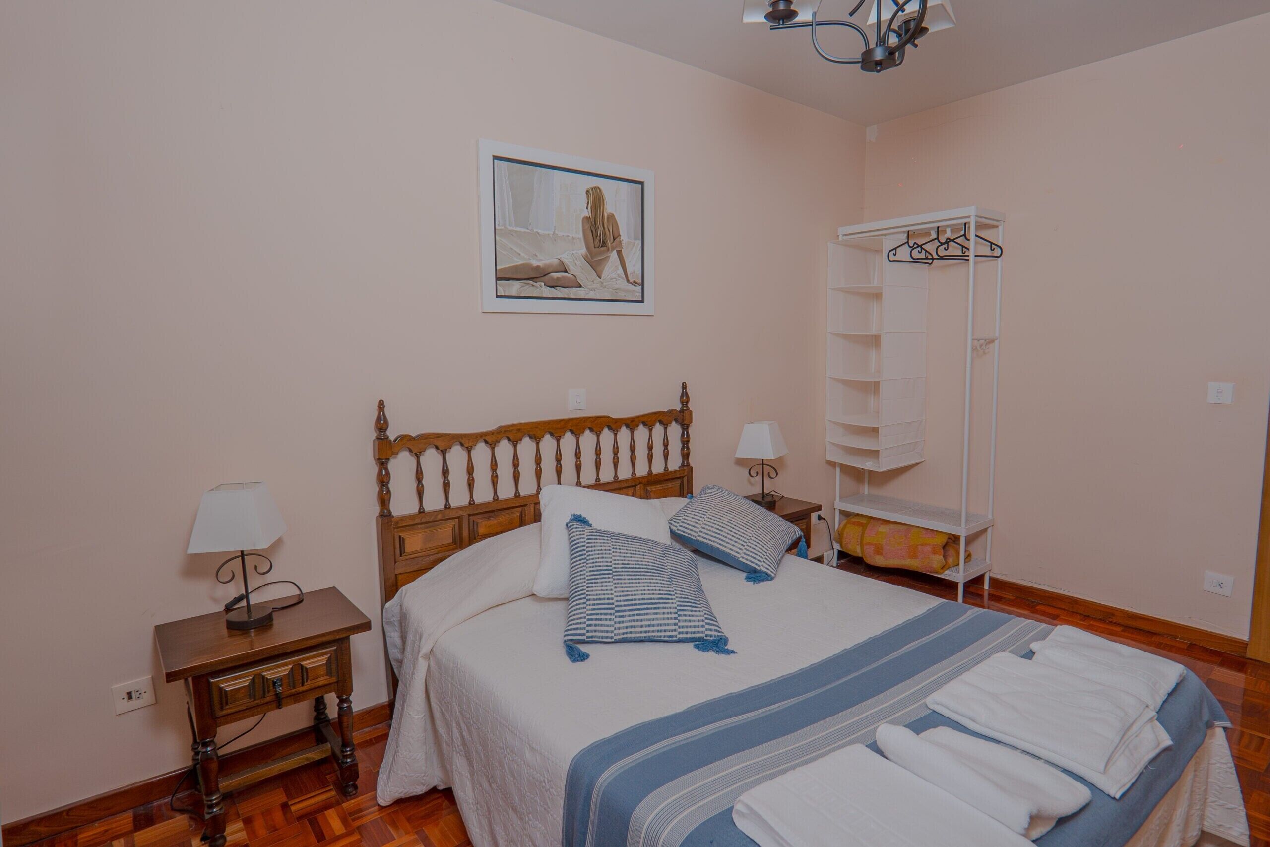 A bedroom with a wooden bed, a bedside table with a lamp, a hanging coat rack, and a framed picture on the wall.