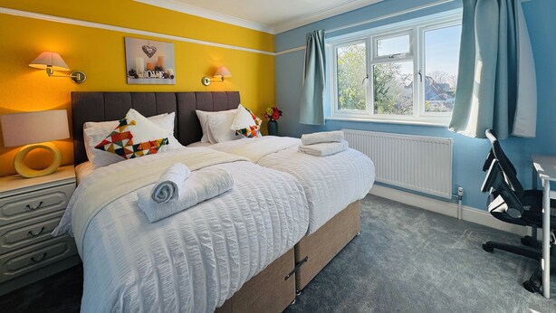 A bedroom with a bed, bedside lamps, a window with curtains, a radiator, and a desk with a chair. - Spacious 4BR house close to Gatwick Airport. (Crawley)