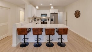 A modern kitchen with a central island, four bar stools, stainless steel appliances, and pendant lighting.