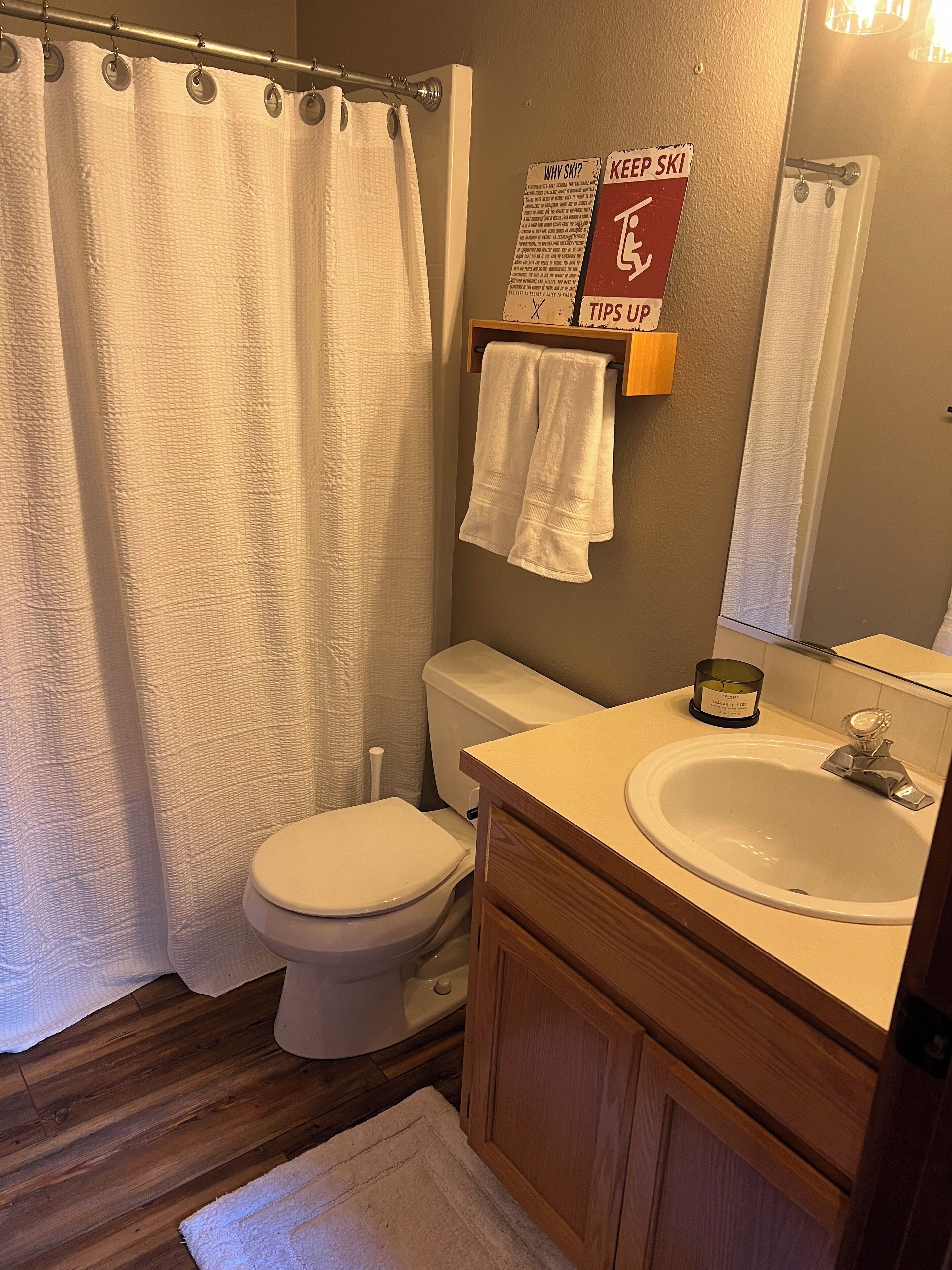 Un baño con inodoro, lavabo y cortina de ducha. Hay un perchero con toallas y un cartel que dice "KEEP SKI TIPS UP".