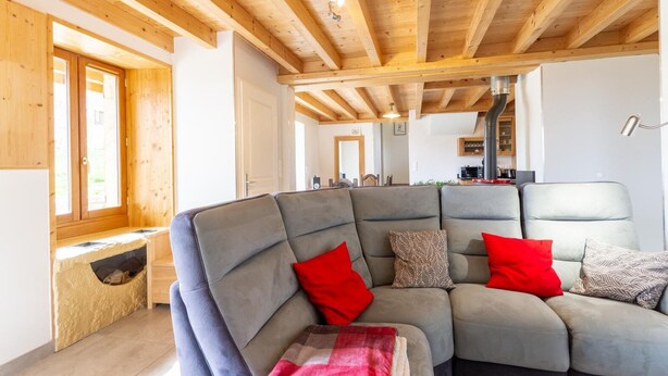 A cozy living room with a sectional sofa, wooden beams, a built-in bench, and a view of the kitchen. - LA MILONGA - AYN - Gîte (AYN)