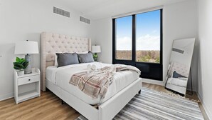 Un dormitorio moderno con un ventanal grande, una cama con cabecera capitonada, mesitas de noche con lámparas y un espejo.