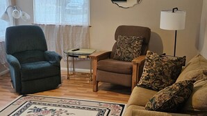 A living room with a blue armchair, a brown armchair with floral cushions, a wooden side table, a floor lamp, and a patterned rug. - The Queen 2 Bedroom Apartment D (Craig)