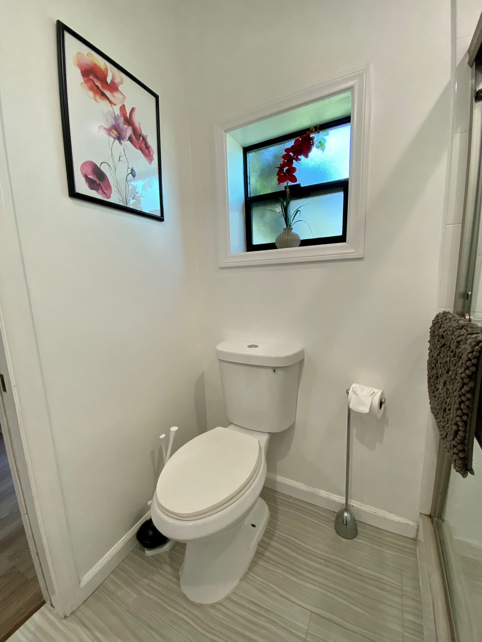 Un baño con inodoro, una ventana pequeña con una planta y una obra de arte floral enmarcada en la pared.