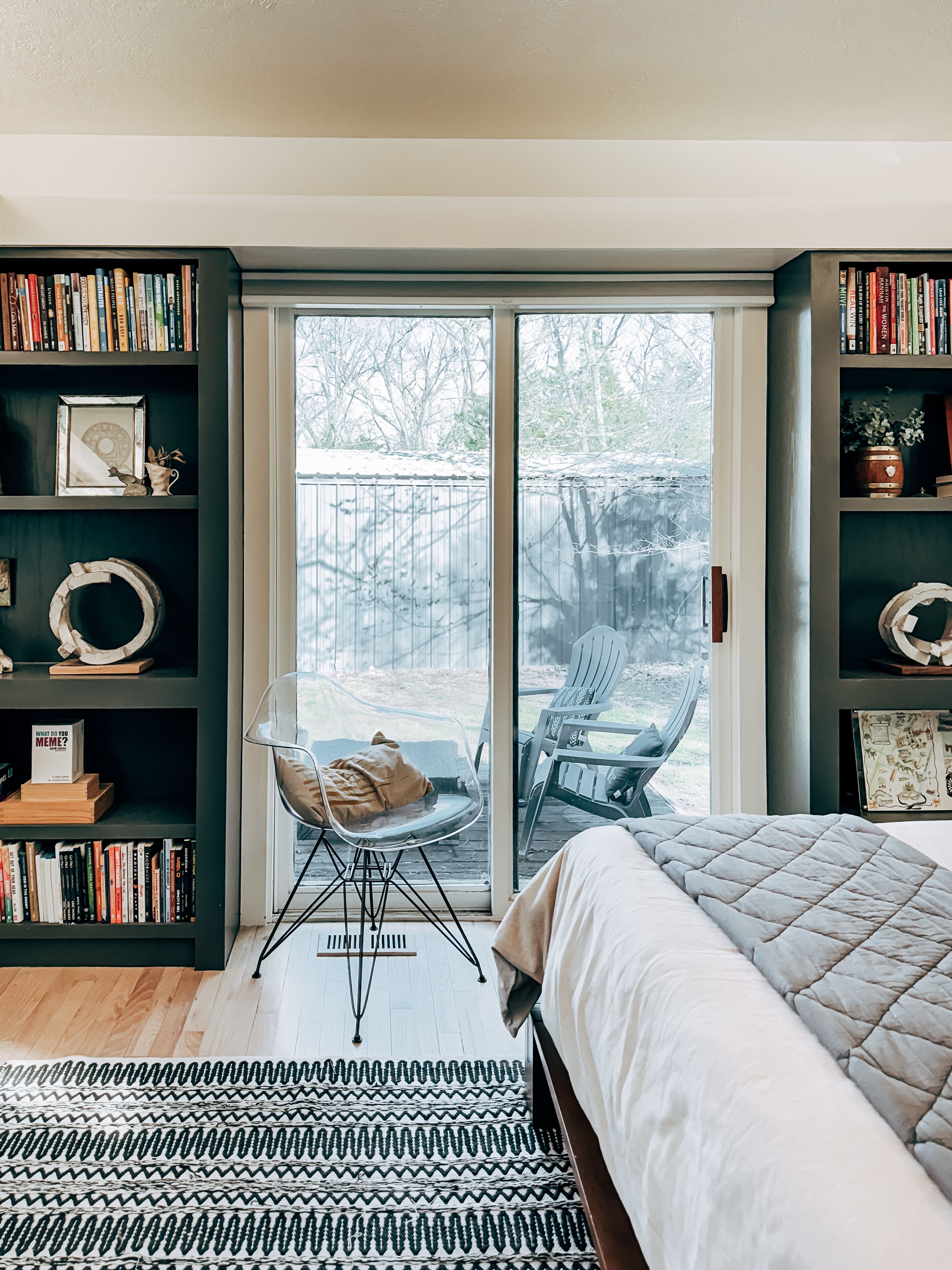 Een slaapkamer met een bed, een groot raam, een schuifdeur naar een terras en twee boekenplanken vol boeken.
