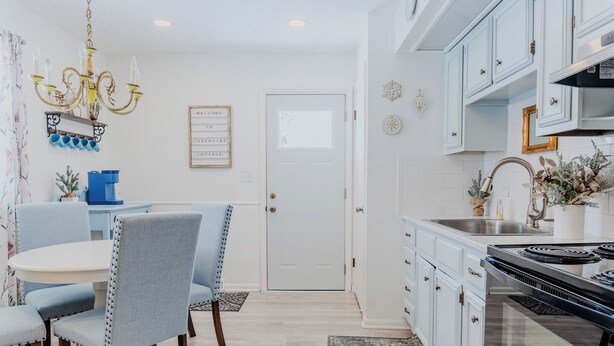 A modern kitchen with white cabinets, a black stove, and a dining area with blue chairs. - Charming Creekside Cottage [] Near Downtown, OSU & Airport (Gahanna)