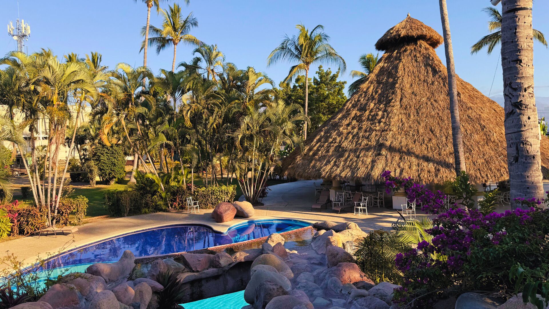 Een resort met een zwembad, palmbomen en een gebouw met rieten dak.
