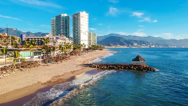 A beach with a pier, beachfront buildings, and mountains in the background.