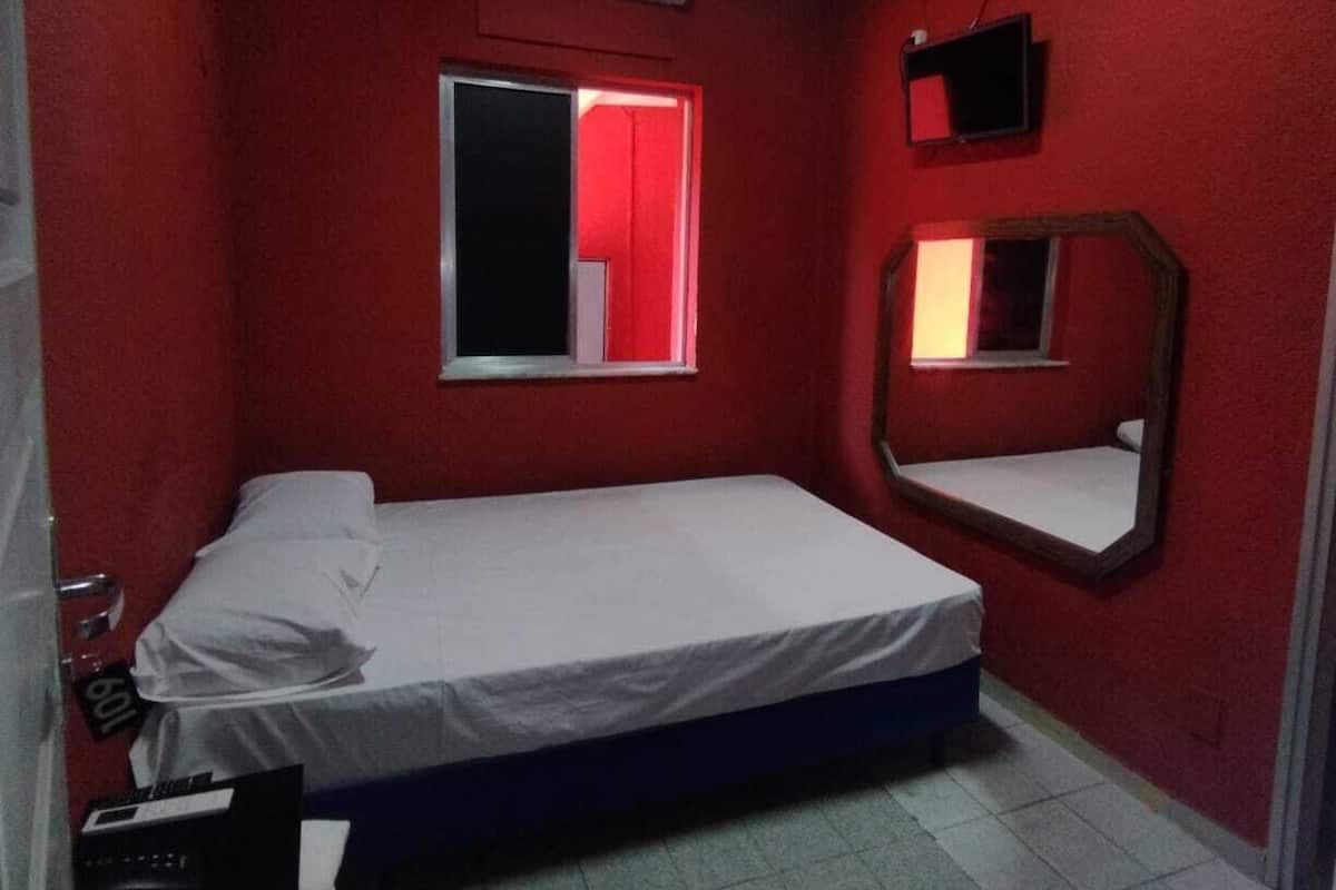 A small, red-walled bedroom with a single bed, a window with red curtains, a wall-mounted TV, and a mirror.