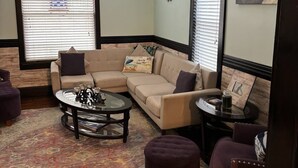 A living room with a beige sectional sofa, a glass coffee table, a purple armchair, and a round side table. There is a ceiling fan, a wall clock, and a framed picture on the wall.